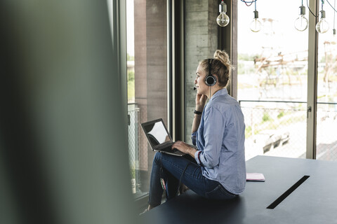 Young businesswoman sitting at desk, making a call, using headset and laptop stock photo