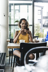 Junge Frau sitzt in einem Café, trinkt Kaffee und liest Textnachrichten - UUF14247