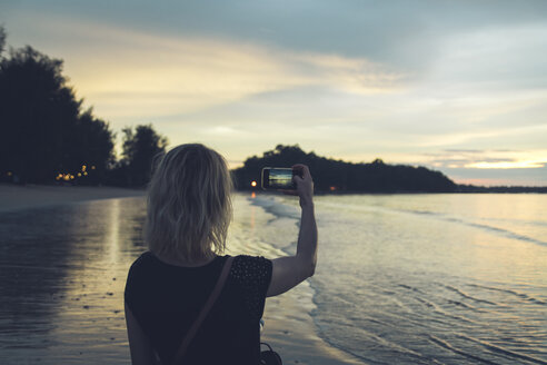 Thailand, Khao Lak, Rückenansicht einer Frau, die am Strand bei Sonnenuntergang mit dem Handy fotografiert - CHPF00481