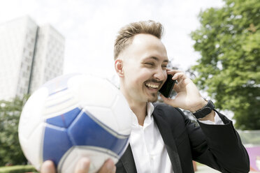 Glücklicher junger Geschäftsmann mit Fußball im Gespräch am Handy in der Stadt - KMKF00381