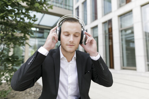 Young businessman with closed eyes wearing headphones outdoors - KMKF00387