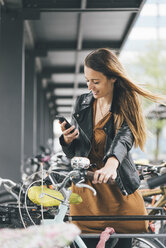 Lächelnde junge Frau mit Fahrrad und Mobiltelefon in der Stadt - KNSF03988