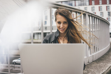 Porträt einer lächelnden jungen Frau mit zerzaustem Haar, die auf einer Autobahnbrücke einen Laptop benutzt - KNSF03999