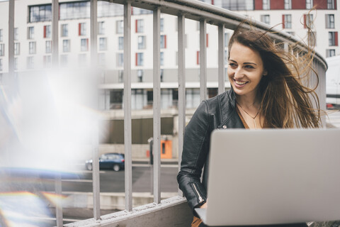 Glückliche junge Frau mit zerzaustem Haar, die auf einer Autobahnbrücke einen Laptop benutzt, lizenzfreies Stockfoto