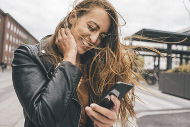 Happy young woman with windswept hair using cell phone in the city - KNSF04005