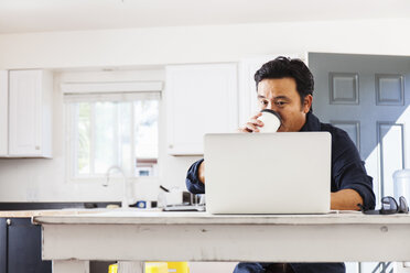 Älterer Geschäftsmann tippt auf einem Laptop, während er am Küchentisch Kaffee zum Mitnehmen trinkt - ISF12790