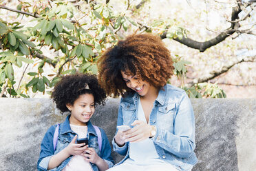 Mutter und Tochter sitzen Seite an Seite und schauen lächelnd auf ihre Smartphones - ISF13070