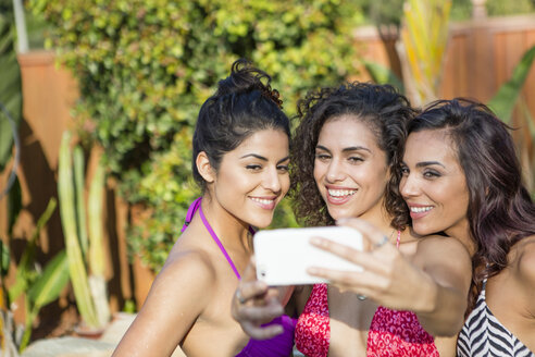 Drei erwachsene Schwestern in Bikinioberteilen posieren für ein Smartphone-Selfie im Garten - ISF13215