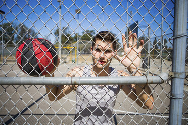 Portrait of young male basketball player behind basketball court fence - ISF13295