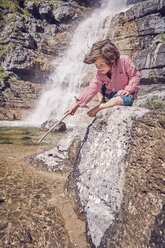 Junge sitzt auf einem Felsen neben einem Wasserfall, hält einen Stock und schaut ins Wasser - ISF13750