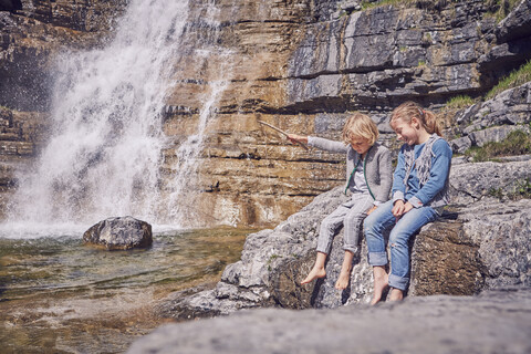 Bruder und Schwester, sitzend auf einem Felsen, entspannend, neben einem Wasserfall, lizenzfreies Stockfoto