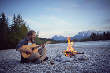 Young man sitting by campfire playing guitar, singing, Wallgau, Bavaria, Germany - ISF13836