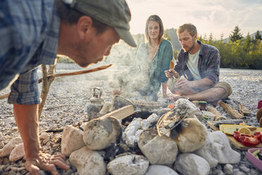 Adults sitting around campfire preparing food and blowing on embers - ISF13845