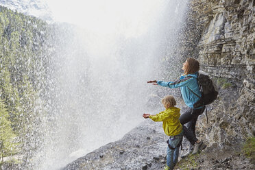 Mutter und Sohn stehen unter einem Wasserfall und strecken die Hände aus, um das Wasser zu spüren, Rückansicht - ISF13974