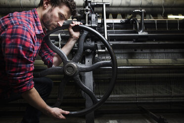 Männlicher Weber dreht das Rad an einer alten Webmaschine in einer Textilfabrik - ISF14086