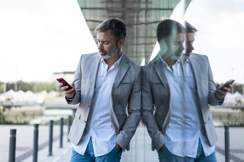 Geschäftsmann lehnt mit Smartphone an einer Glasfassade, lizenzfreies Stockfoto