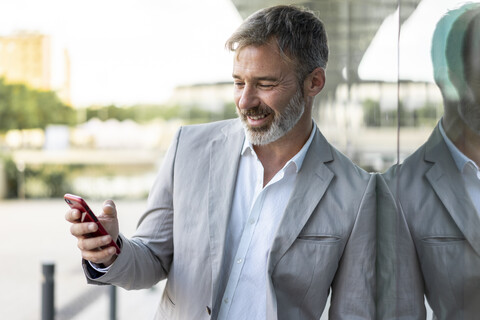 Porträt eines lächelnden Geschäftsmannes, der an einer Glasfassade lehnt und auf sein Smartphone schaut, lizenzfreies Stockfoto