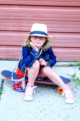 Portrait of young girl wearing tutu, denim jacket and hat, sitting on skateboard - ISF14153