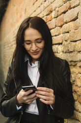Lächelnde junge Frau, die ein Handy an einer Backsteinmauer benutzt - ALBF00568