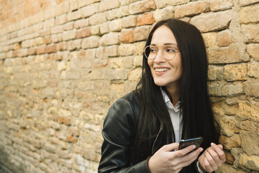 Lächelnde junge Frau mit Handy an einer Backsteinmauer, die sich umschaut - ALBF00569