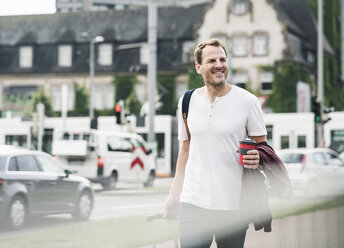 Lächelnder Mann mit Rollkoffer und Kaffee zum Mitnehmen beim Spaziergang in der Stadt - UUF14267