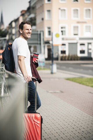 Lächelnder Mann mit Rollkoffer und Kaffee zum Mitnehmen in der Stadt, lizenzfreies Stockfoto