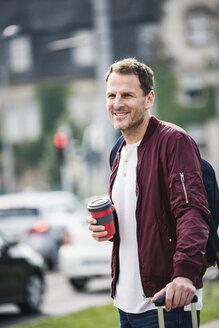 Lächelnder Mann mit Rollkoffer und Kaffee zum Mitnehmen in der Stadt - UUF14271