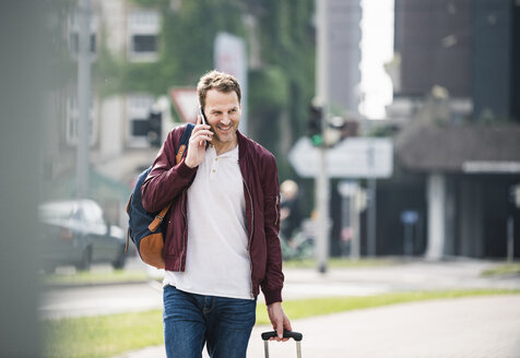 Lächelnder Mann mit Rollkoffer am Handy in der Stadt - UUF14273