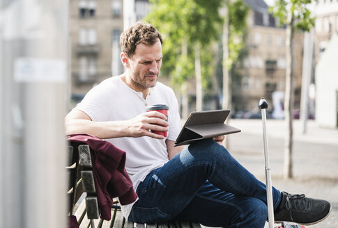 Mann mit Rollkoffer und Kaffee zum Mitnehmen sitzt auf einer Bank und benutzt ein Tablet - UUF14274