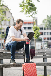 Lächelnder Mann mit Rollkoffer und Kaffee zum Mitnehmen sitzt auf einer Bank und benutzt ein Tablet - UUF14279