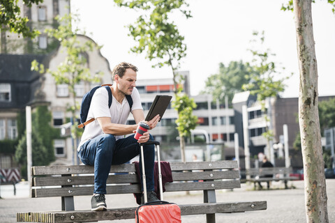 Mann mit Rollkoffer und Kaffee zum Mitnehmen sitzt auf einer Bank und benutzt ein Tablet, lizenzfreies Stockfoto