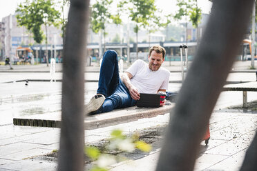 Smiling man lying on bench using tablet - UUF14286