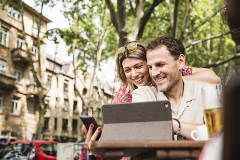 Lächelndes Paar teilt sich Tablet und Handy in einem Café im Freien - UUF14313