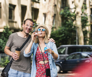 Happy couple with coffee to go blowing soap bubbles in the city - UUF14320