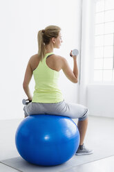 Junge Frau sitzt auf einem Gymnastikball, Rückansicht, hebt Gewichte - CUF34283