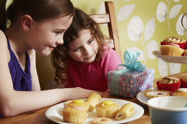 Two girls looking at birthday present with plate of cakes - CUF34388