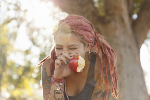 Junge Frau mit rosa Dreadlocks kichert beim Essen eines Apfels im Park - ISF14331