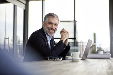 Porträt eines lächelnden Geschäftsmannes mit Laptop am Schreibtisch im Büro, lizenzfreies Stockfoto
