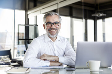 Portrait of smiling businessman with laptop at desk in office - RBF06325