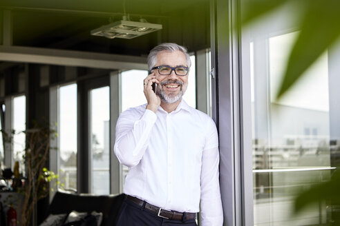 Lächelnder Geschäftsmann, der an einer französischen Tür steht und mit einem Handy spricht - RBF06332