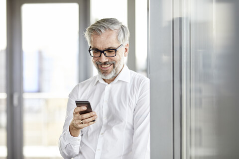Lächelnder Geschäftsmann am Fenster, der auf sein Handy schaut, lizenzfreies Stockfoto