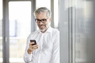 Lächelnder Geschäftsmann am Fenster, der auf sein Handy schaut - RBF06342