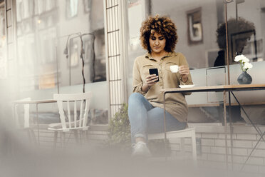 Woman in front of coffee shop, drinking coffee, holding smart phone - KNSF04047