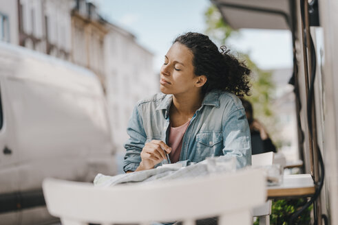 Junge Frau sitzt vor einem Café mit geschlossenen Augen und genießt die Sonne - KNSF04096