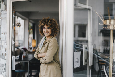 Smiling woman standing with arms crossed in door of coworking space - KNSF04155