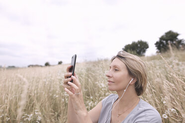 Frau mit Kopfhörern auf einem Feld nimmt Selfie mit Smartphone - KMKF00392