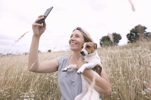 Frau mit Hund macht Selfie mit Smartphone auf einem Feld, lizenzfreies Stockfoto