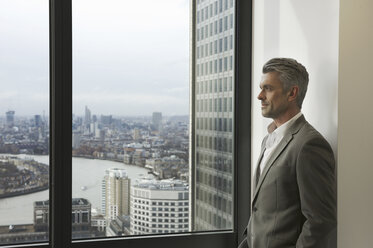 Portrait of mature businessman looking out of office window, Canary Wharf, London, UK - CUF34586