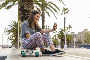 Glückliches Teenager-Mädchen, das auf einem Skateboard sitzt und mit Kopfhörern Musik hört - WPEF00466