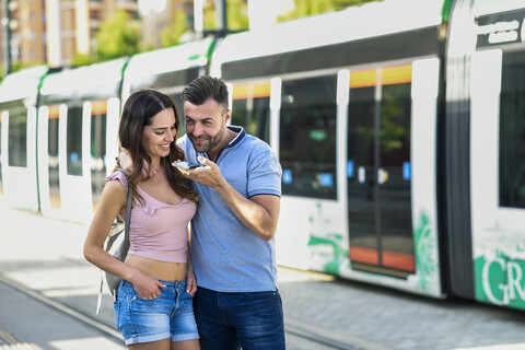 Ein Paar benutzt sein Smartphone, während es am Bahnhof auf die Straßenbahn wartet, lizenzfreies Stockfoto